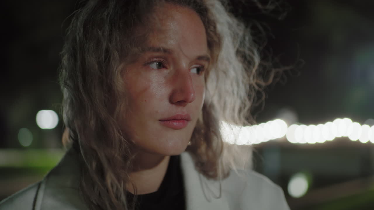 Pensive young woman with loose hair swaying in gentle wind gazes into distance under night sky, surrounded by blurred bokeh lights and calm atmosphere near water, lost in deep personal thought