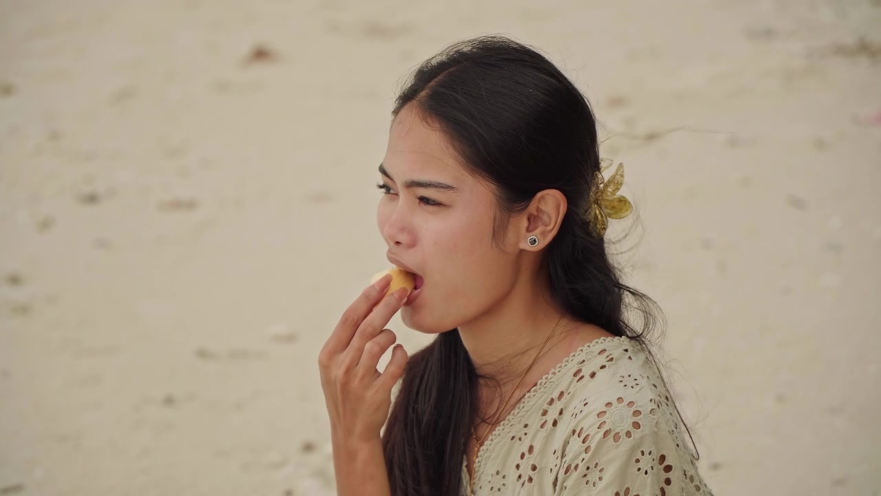 砂浜でのクローズアップポートレート。表情豊かな口元と目を捉え、耳に挟んだ花、ゆるく流れる黒髪、柔らかな光が特徴的。微かな呼吸と声の表現が、自然な演技のような感情豊かな雰囲気を醸し出す。
