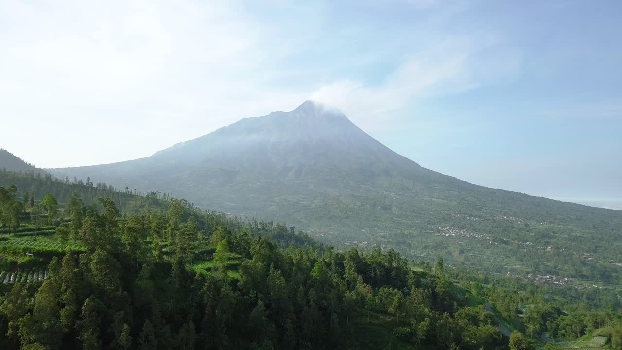 Gunung Merapi dengan pemandangan pedesaan perkebunan sayur dan pepohonan lebat dalam cuaca sedikit berkabut, Jawa Tengah, Indonesia. Pemandangan udara 4K perkebunan dengan pemandangan pedesaan Jawa Tengah