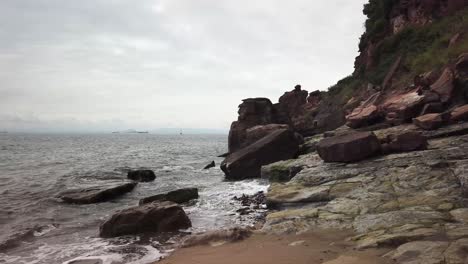 Welle-Bricht-Auf-Felsen-An-Einem-Strand-In-West-Wemyss,-Fife,-Schottland,-Mit-Segelboottankern-Und-Edinburgh-In-Der-Ferne