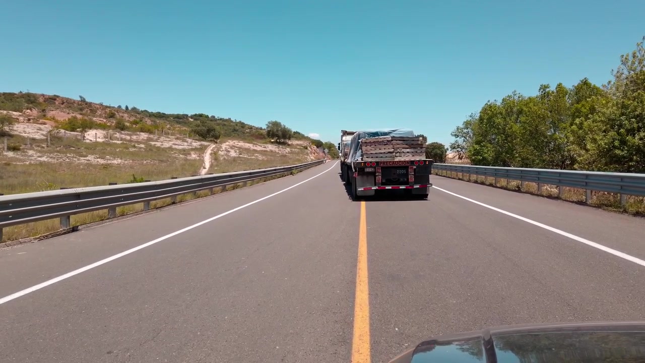 POV di guida che mostra camion in viaggio lungo l'autostrada con guardrail, terreno roccioso e vegetazione verde vicino a Huatulco, Oaxaca, Messico, sotto un cielo azzurro e limpido durante il giorno