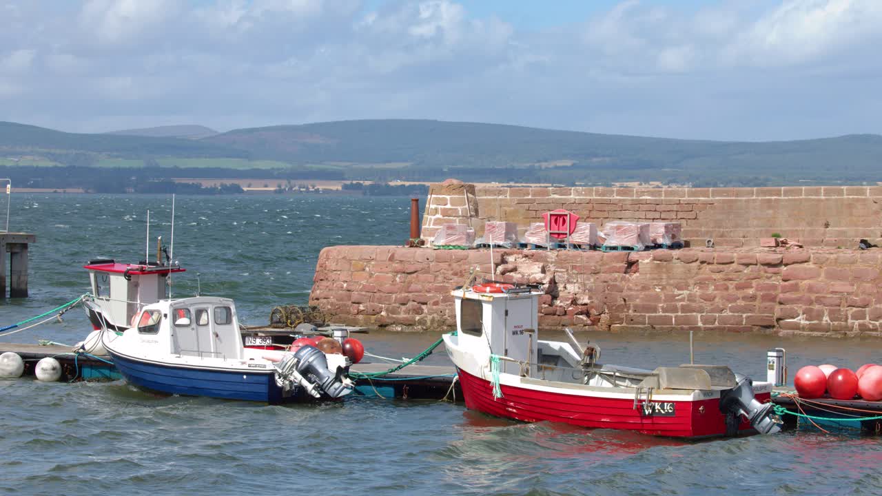 Two fishing boats gently bob in Cromarty harbour, bright daylight, slight camera pan, coastal backdrop
