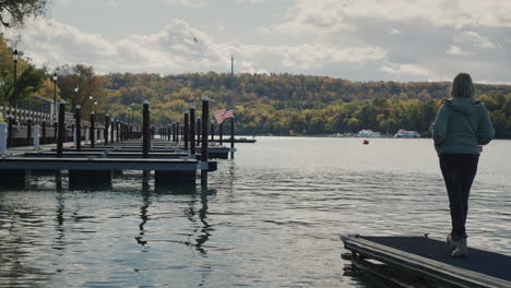 Eine-Frau-Steht-Auf-Einem-Pier-Mit-Blick-Auf-Den-Ontariosee,-Die-Amerikanische-Flagge-Ist-In-Der-Ferne-Zu-Sehen,-Es-Sind-Keine-Anderen-Menschen-Und-Yachten-In-Der-Nähe.-Herbst-Und-Ende-Der-Touristensaison.