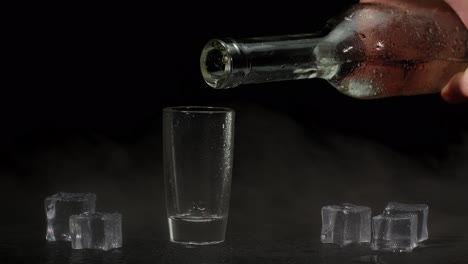 Barman-hand-pours-vodka,-tequila-or-sake-from-bottle-into-glass-on-black-background-with-ice-cubes