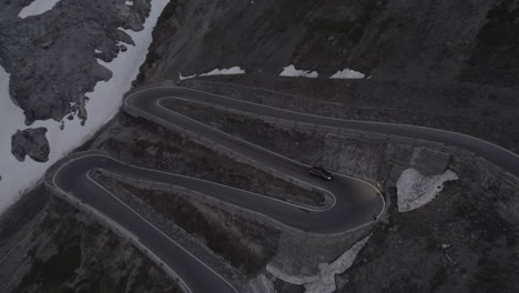 Toma-De-Drone-De-Un-Automóvil-Conduciendo-Cuesta-Abajo-En-El-Paso-Stelvio-Italia-En-Un-Día-Gris-Con-Nieve-En-Las-Montañas-En-Una-Carretera-Sinuosa-Y-Faros-Encendidos-Durante-El-Atardecer