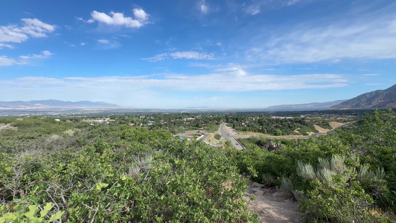 Una vista elevada amplia sobre el Valle de Salt Lake en Utah, mirando a través de las verdes estribaciones hacia la expansión urbana y las montañas distantes. La escena encaja con la amplia cuenca del área de Salt Lake City