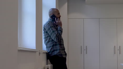 an asian indian male casually walking up to a window seeking privacy to take a telephone call on his mobile phone in an office environment