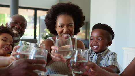 Mehrgenerationenfamilie-Sitzt-Zu-Hause-Am-Tisch-Und-Jubelt-Mit-Wasser-In-Richtung-Kamera