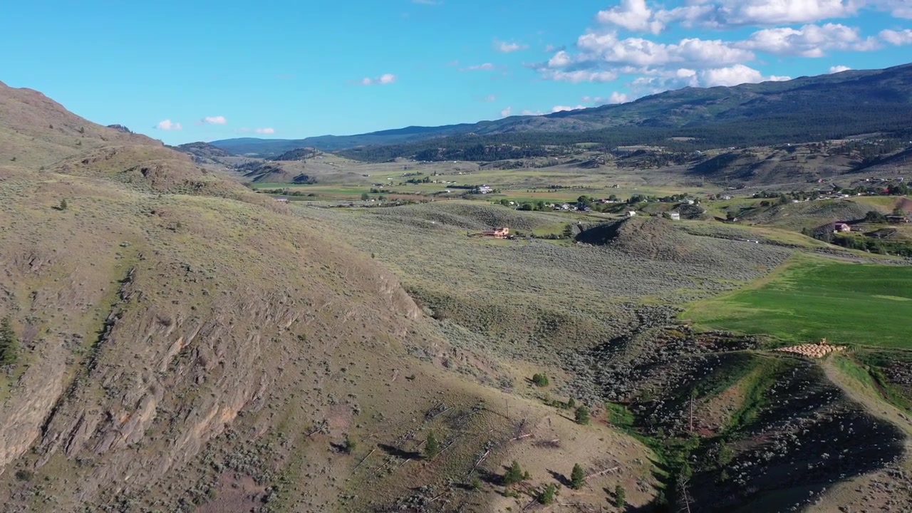 Ripresa aerea con drone che domina un vasto lago calmo incastonato tra montagne aspre e aride. La luminosa luce del sole estivo illumina i pendii erbosi e il cielo azzurro limpido della natura selvaggia dell'interno della Columbia Britannica.