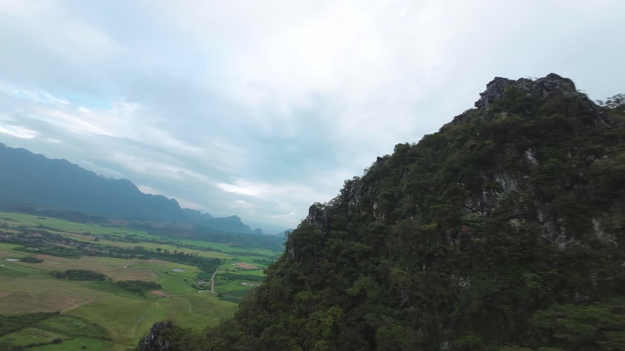 Drone melayang di atas Laguna Biru, Laos, dengan lembah hijau dan tebing di bawah langit mendung yang muram