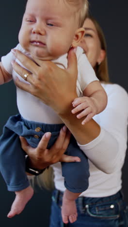blonde long-haired woman wearing white shirt and jeans holding a baby. mother waves her baby and kid starts to cry. black backdrop. vertical video