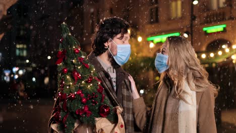 caucasian happy couple wearing facial masks holding christmas tree and talking while snowing on the street in christmas