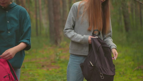 close-up of siblings walking through a peaceful forest, each carrying backpacks, they pause to drop their bags, and one begins to open hers to retrieve an item, surrounded by lush greenery