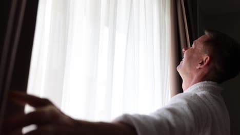 Man in bathrobe stretches and opens curtains in a hotel room