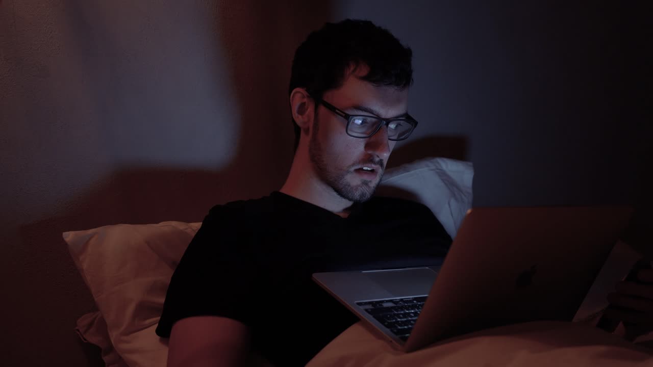 Man with laptop in bed, online internet at night, late work web browsing video watching on notebook before sleep