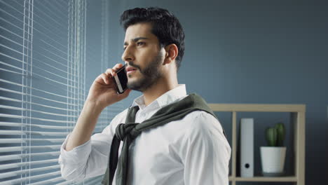 close up view of arabian businessman standing near the window in his office and speaking on the phone, then looking out the window