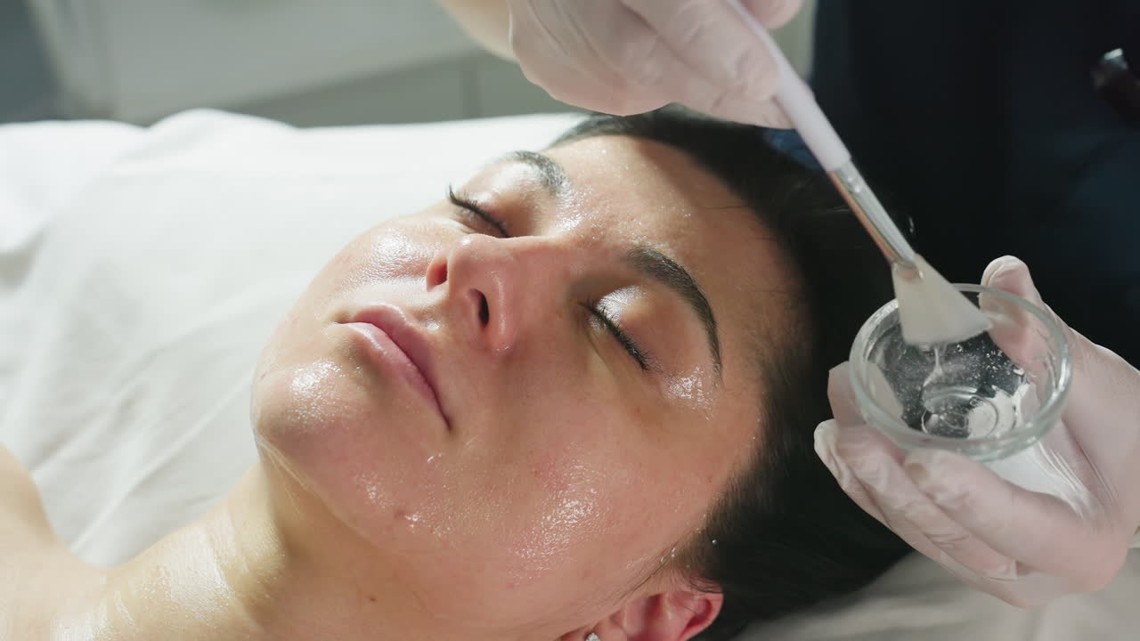 Beautiful lady lies with eyes closed on spa bed while facialist wearing gloves dips brush into clear gel and smoothly applies nourishing mask in gentle strokes for radiant skin treatment