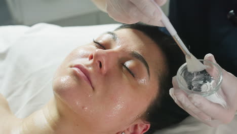 beautiful lady lies with eyes closed on spa bed while facialist wearing gloves dips brush into clear gel and smoothly applies nourishing mask in gentle strokes for radiant skin treatment