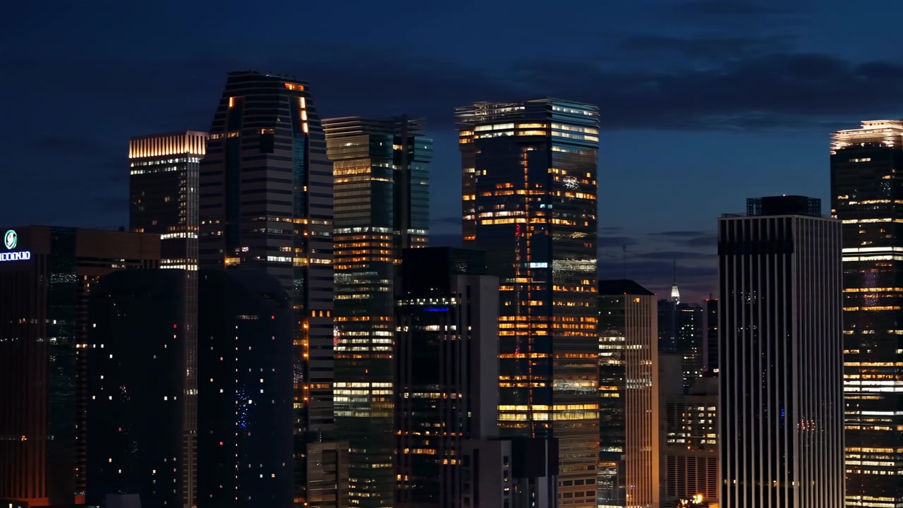 Edifícios altos deslizam gradualmente para a direita enquanto a câmera se move para a esquerda através da densa arquitetura do centro da cidade, com janelas brilhantes, fachadas refletivas e um céu azul escuro ao entardecer