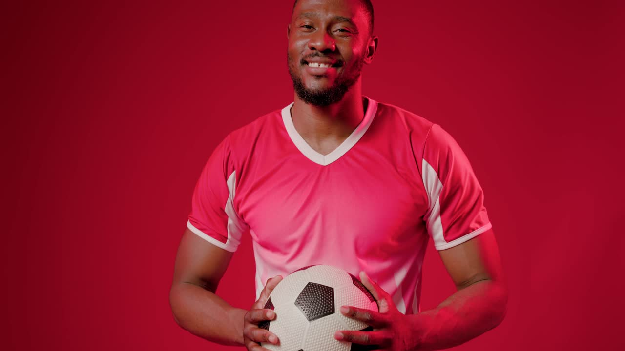 Hombre africano reaccionando a la señal de la cámara, sosteniendo un balón de fútbol, sonriendo y luego calmándose para la sesión con la camiseta roja.