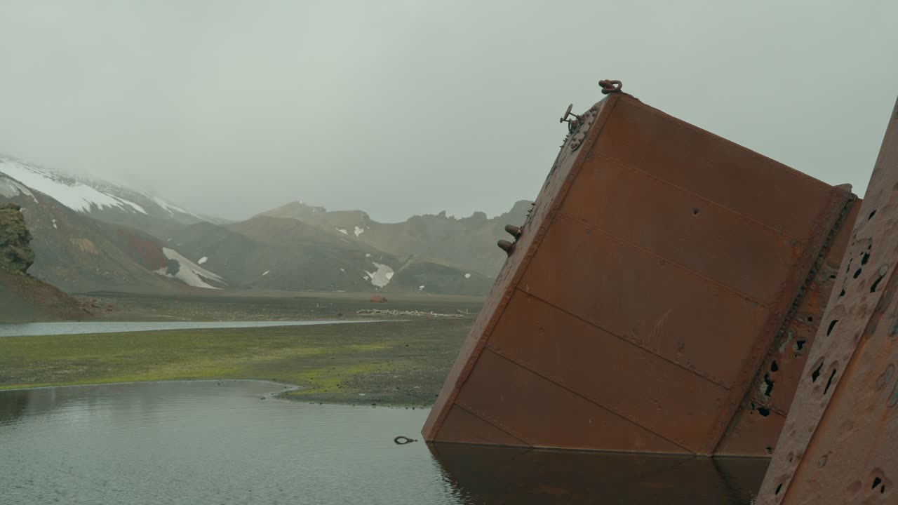 Massiv rusten industritank fra historisk hvalfangststation tipper ned i lagunen ved Whalers Bay, Deception Island, Antarktis, med vulkanske, sneplettede bjerge i baggrunden.