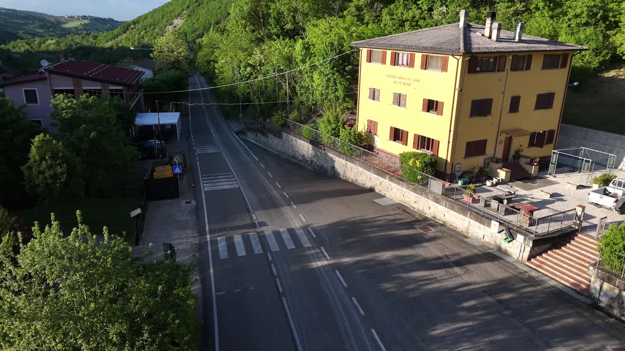 Descending drone shot over residential street with yellow house and surrounding forested hills near Mignano Dam, clear road lines and distant terrain visible under daylight, real time