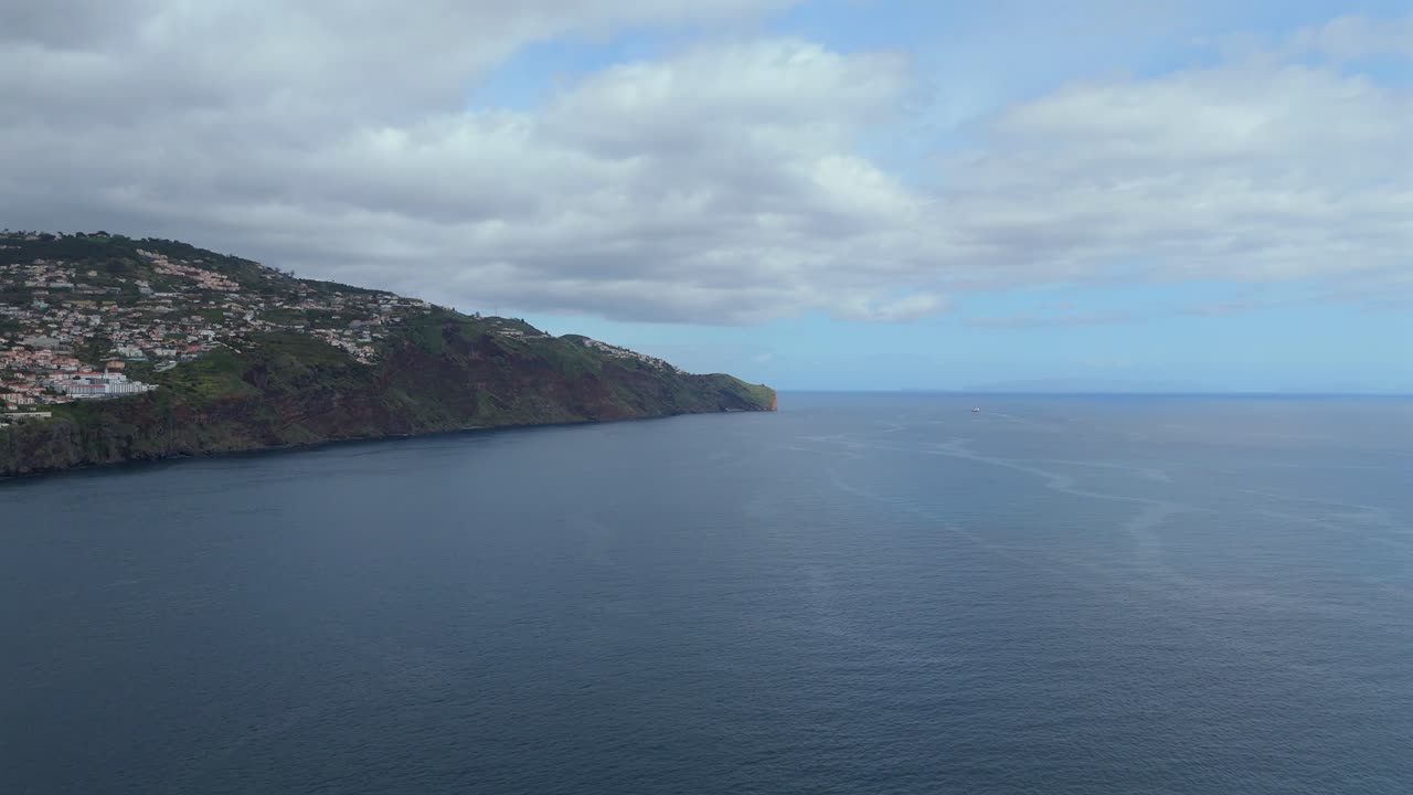 Filmato aereo che mostra la spettacolare costa di Madeira, Portogallo, con scogliere a picco sull'oceano, case distanti e un vasto paesaggio marino atlantico sotto cieli nuvolosi