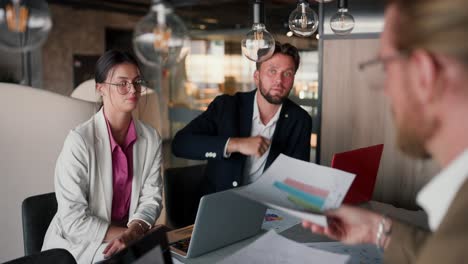 a brunette man in a blue jacket offers his solution to a problem on paper in the office. a trio of office workers sitting at a table and looking at their task schedule