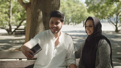 young muslim couple sitting on bench in park, taking selfie.