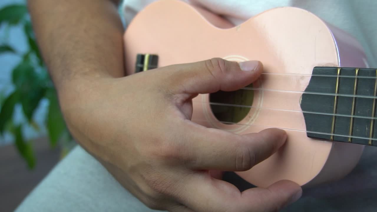 hombre de mediana edad 30 y con cuentas está jugando y silbando con su ukulele rosado al lado de una gran ventana y cerca de una planta verde
