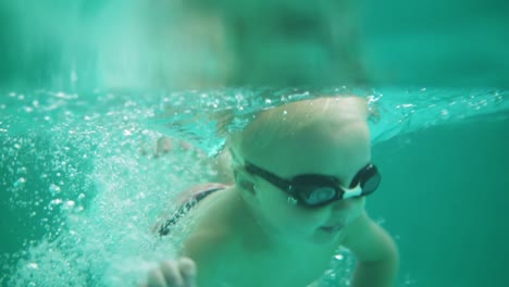 Eine-Unterwasseraufnahme-Eines-Süßen-Blonden-Kleinkindes-Mit-Schutzbrille,-Das-Im-Schwimmbad-Unter-Wasser-Schwimmt