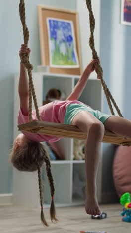 little girl with long hair in plaits lies on back on swing near window in room. playful kid in pink t-shirt and turquoise shorts relaxes at home