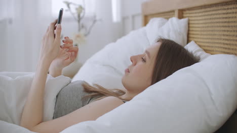 woman in morning sitting in bedroom leaning against the soft leather back of bed. write messages with your smartphone use apps to information