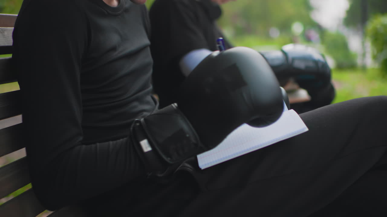 boxer gloves resting on bench, athlete sits outdoors after intense training with black leather gloves and notepad under glove, pen tip visible on pad, companion blurred in background, calm reflective