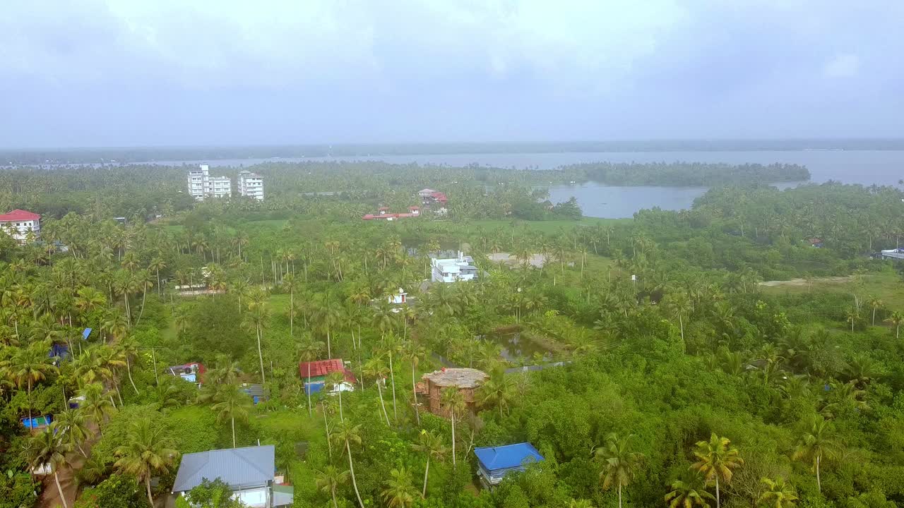 Expansive aerial view of a large, rural building situated between a narrow road and waterlogged tropical marshland paddy fields and dense coconut palms in Kerala, India
