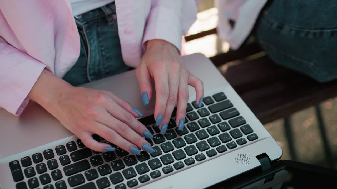 primer plano de manos con uñas pulidas escribiendo en el teclado de la computadora portátil al aire libre, sentado en un banco con una vista parcial de otra persona al lado, los detalles incluyen manicure elegante y trabajo activo