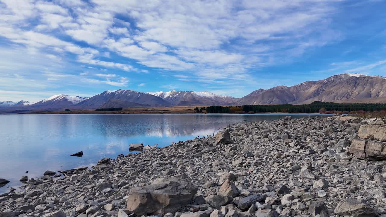 Drone footage captures Lake Tekapo's serene waters and rocky shoreline under a vibrant sky, showcasing the natural beauty of New Zealand