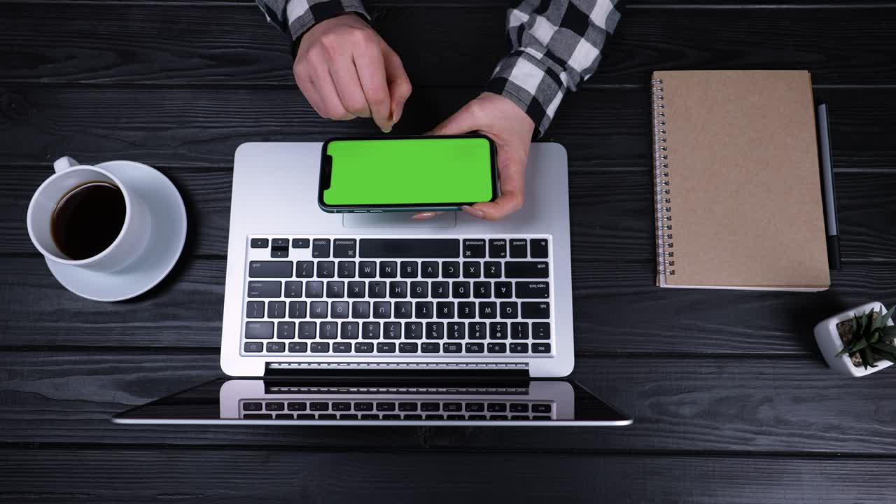 An overhead view of the girl's hands holding a smartphone with a green screen and a chrome key. The girl sits at a table on which there is an open laptop and a cup of coffee. Slow motion. Close up