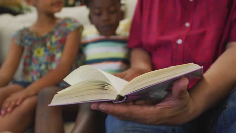 Mid-section-of-grandfather-and-grandchildren-reading-book-at-home