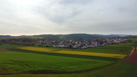 Deutsches-Dorf-Auf-Der-Ebene-Inmitten-Grüner-Felder-Und-Sanfter-Hügel,-Niedrige-Drohnenansicht