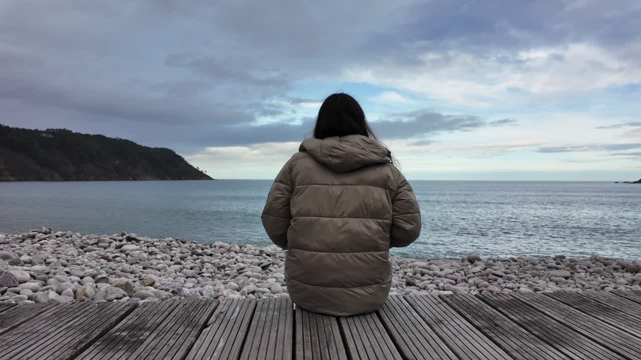 Slow-motion shot of a woman sitting alone on a wooden deck facing the waves