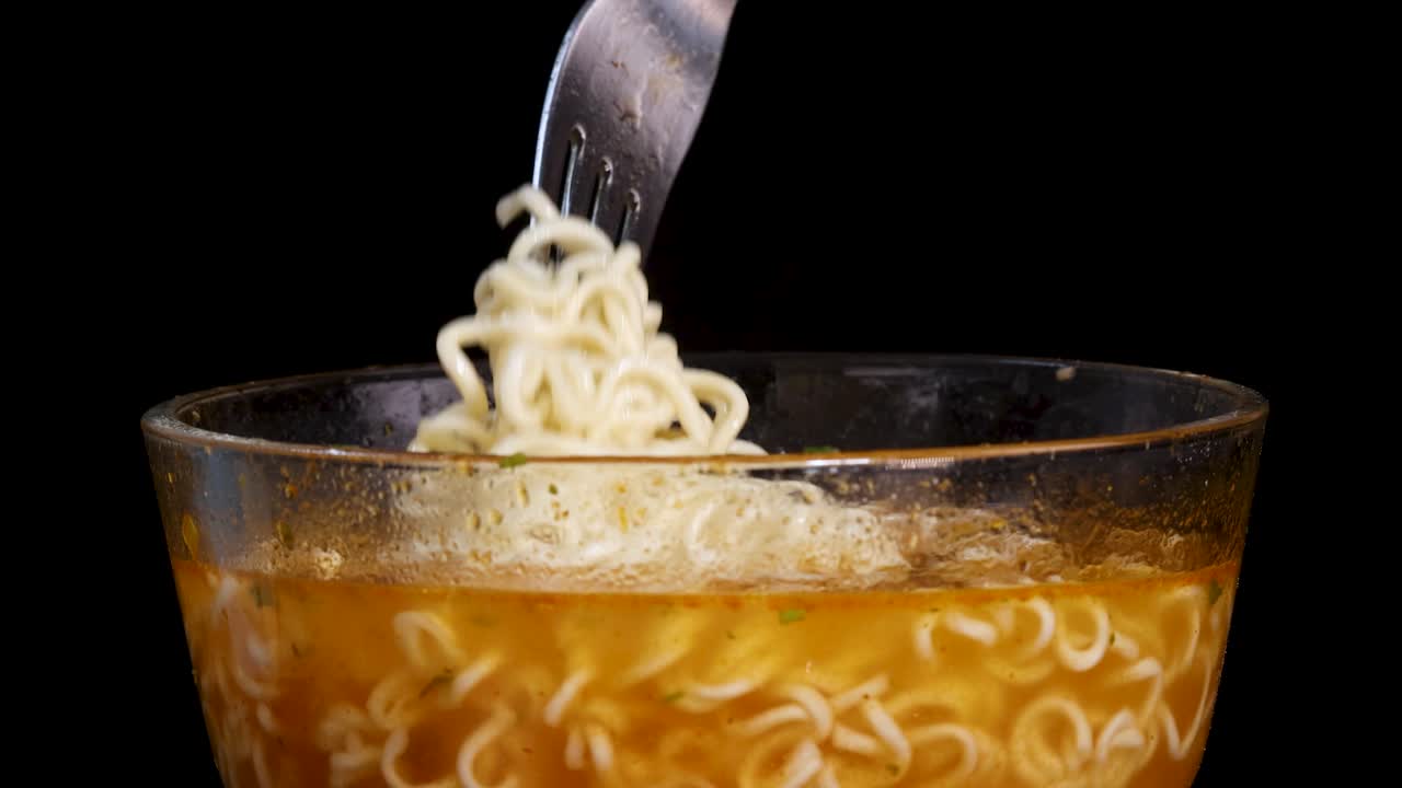 A hand uses a fork to lift cooked instant noodles from a clear glass bowl filled with broth, under bright studio lighting against a black background