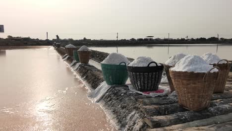 Cestas-Llenas-De-Sal-Marina-Natural-Recolectada-Del-Lago-Salado-En-Tailandia-Con-Un-Granjero-Recogiendo-Sal-En-El-Fondo-Contra-La-Cálida-Luz-Del-Sol