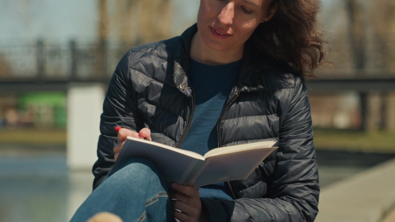 Close up of woman writing in open notebook with red pen while seated outdoors on sunny day. Notebook pages visible as woman writes wearing dark puffer jacket and blue shirt. Background shows