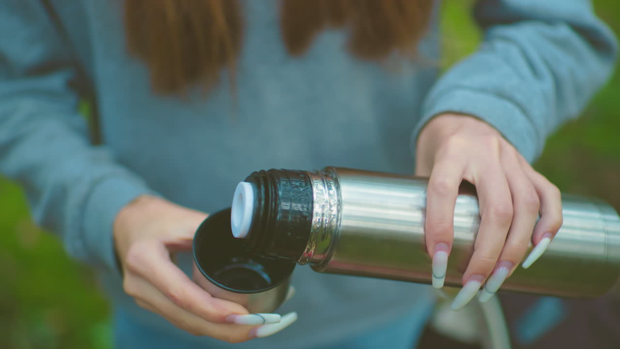 primer plano de alguien con uñas pulidas presionando la tapa del termo para verter agua en la cubierta, en contra de la vegetación borrosa, destacando el manejo cuidadoso y centrarse en la bebida refrescante
