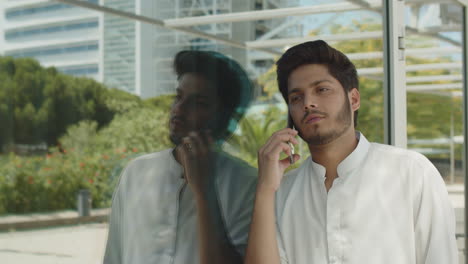 handsome arabic man talking on cellphone, leaning on glass wall.