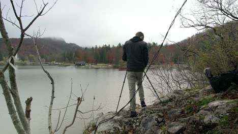 A-man-setting-up-his-tripod-on-a-rock-ledge-surround-by-a-lake-in-the-background-with-trees-in-the-foreground-and-the-sun-shining-in-the-background-during-the-fall-season