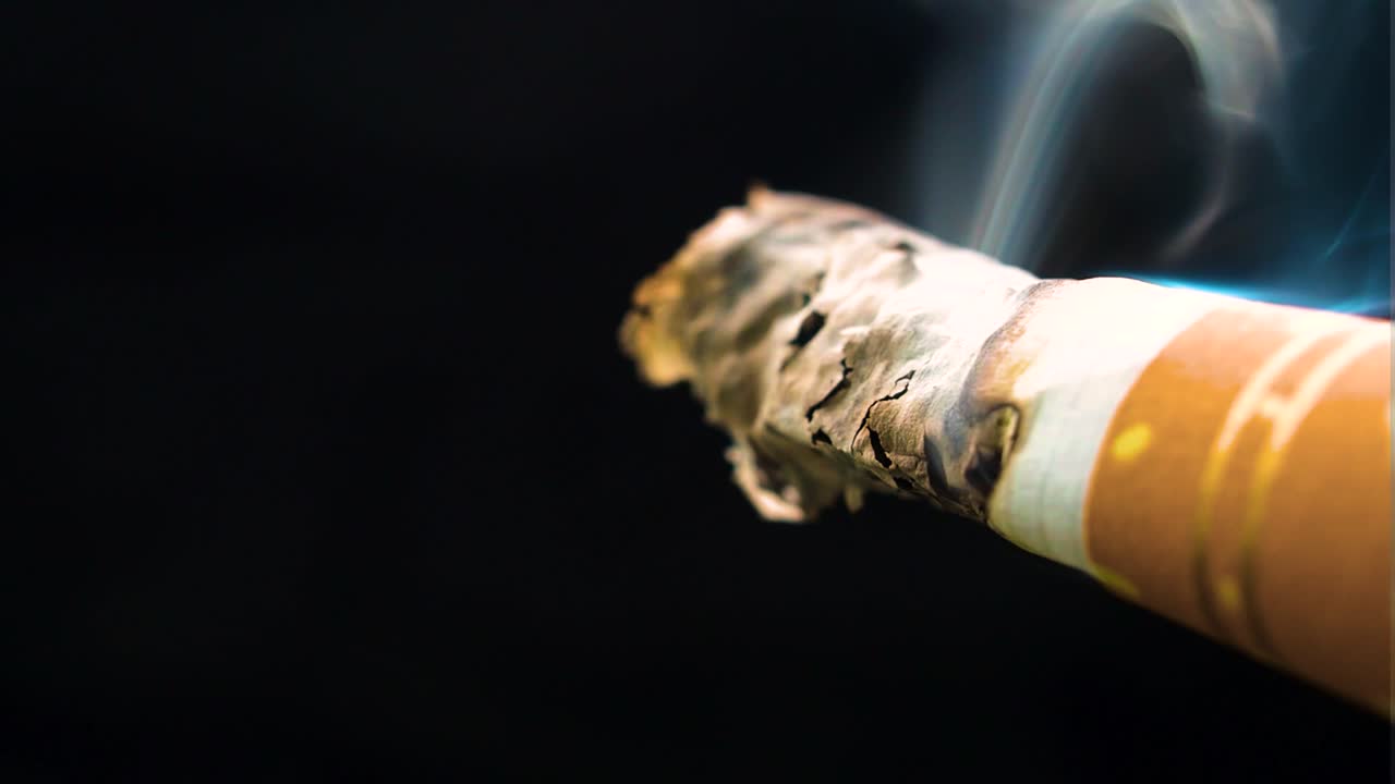 very tiny bit of a cigarette almost at the end of the filter burning clouds of smoke moving in all directions affected by the breeze white paper turning black ash stays golden filter stripes darkness