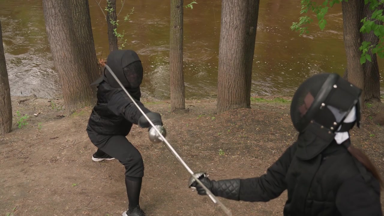 Two women in black fencing gear engage with swords on dirt path beside trees and water demonstrating dynamic movements. Their protective masks cover faces as they assume various stances