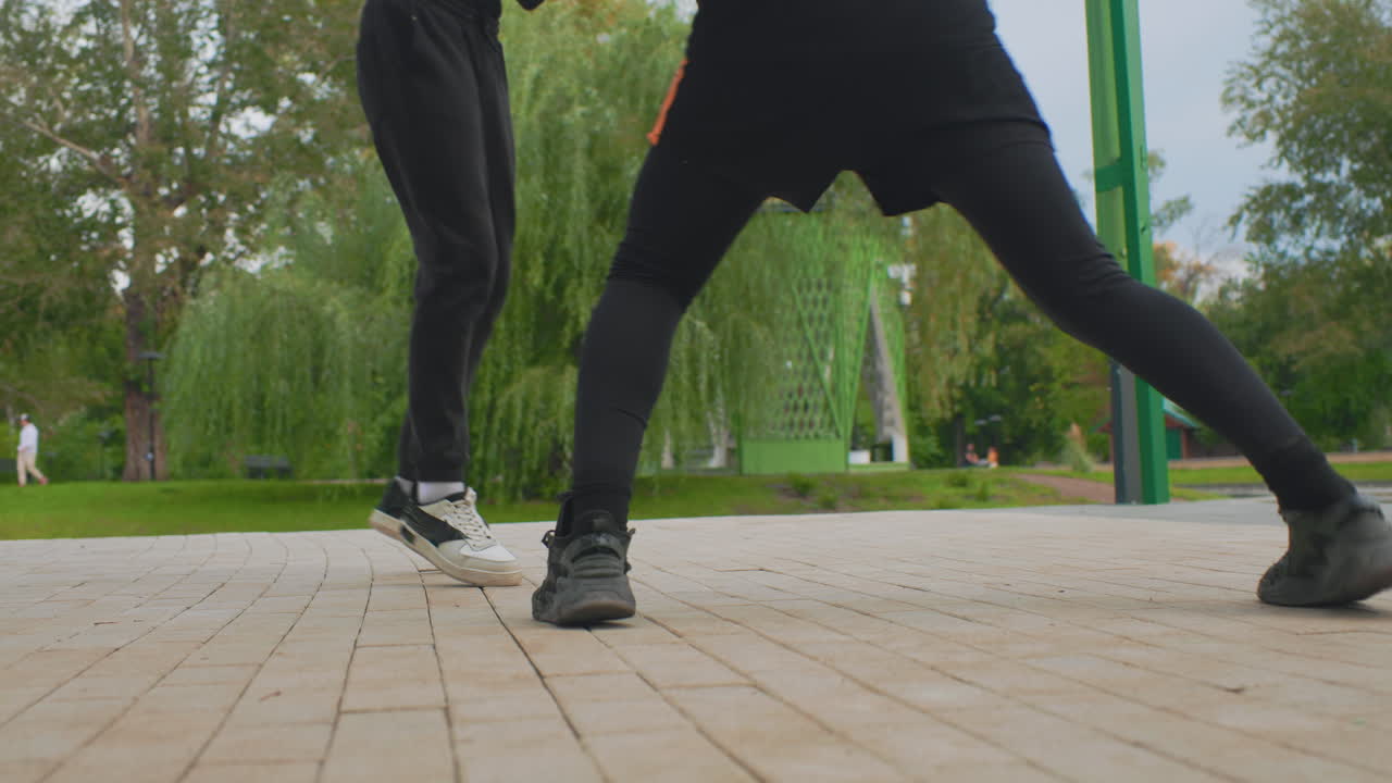 park plaza dynamic footwork training session with synchronized steps and pivots, two athletes in leggings and mixed sneakers rehearse drills and slides on paved promenade, focused coachlike